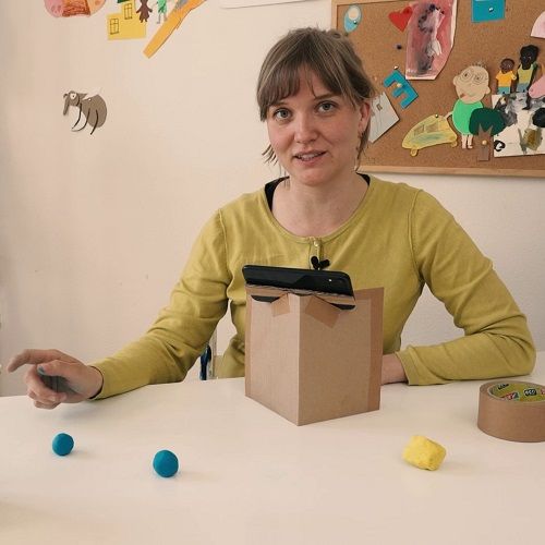 Eine Frau zeigt auf bunte Kugeln aus Knete auf einem Tisch, darüber liegt ein Smartphone.