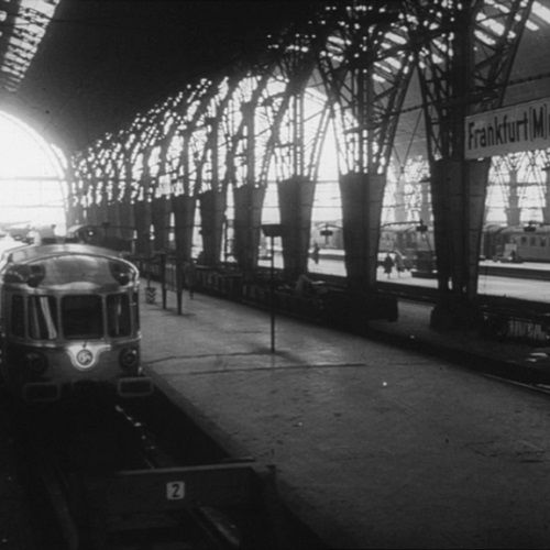 In schwarz-weiß der Frankfurter Hauptbahnhof mit einem Zug, Menschen am Glas und dem Dach.