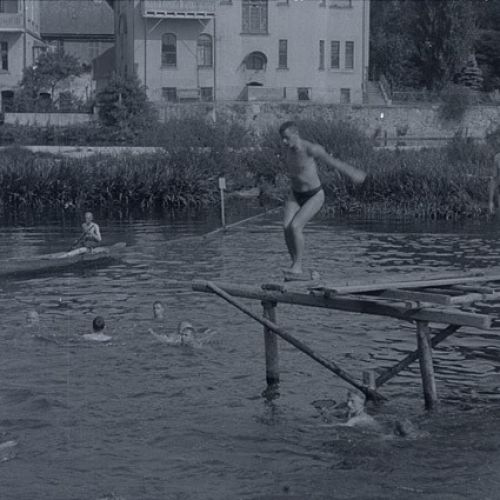 In schwarz-weiß ein Mann der im Freibad von einem Steg ins Wasser springt.