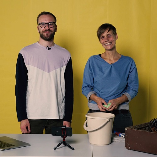 Ein Mann und eine Frau stehen vor einem Tisch mit einem Smartphone, einem Koffer und einem Putzeimer.