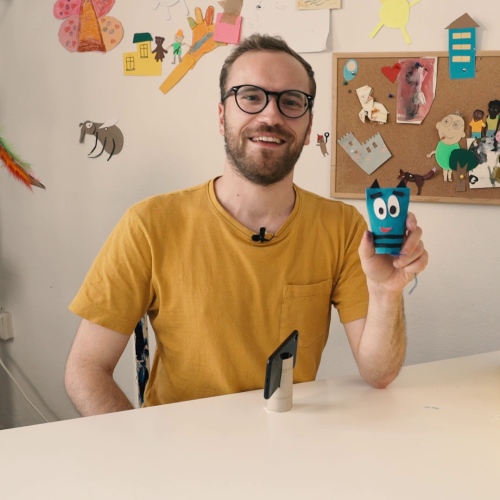Ein Mann lacht und zeigt eine gebastelte Figur mit fröhlichem Gesicht.